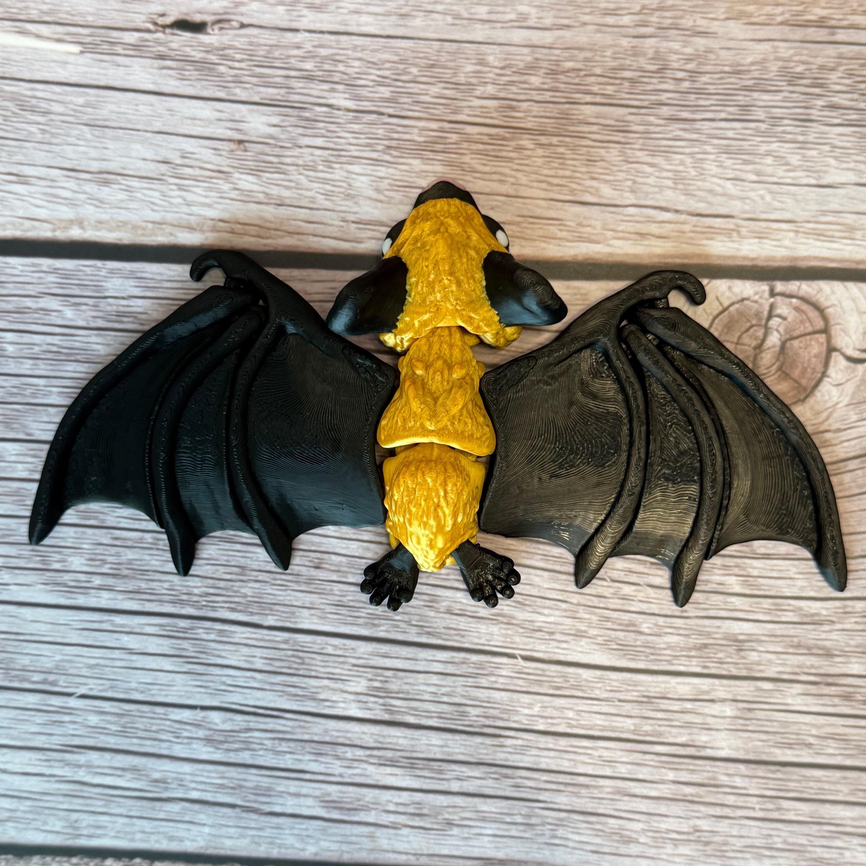 3D printed gold and black articulated bat with pink nose, placed on a wooden surface