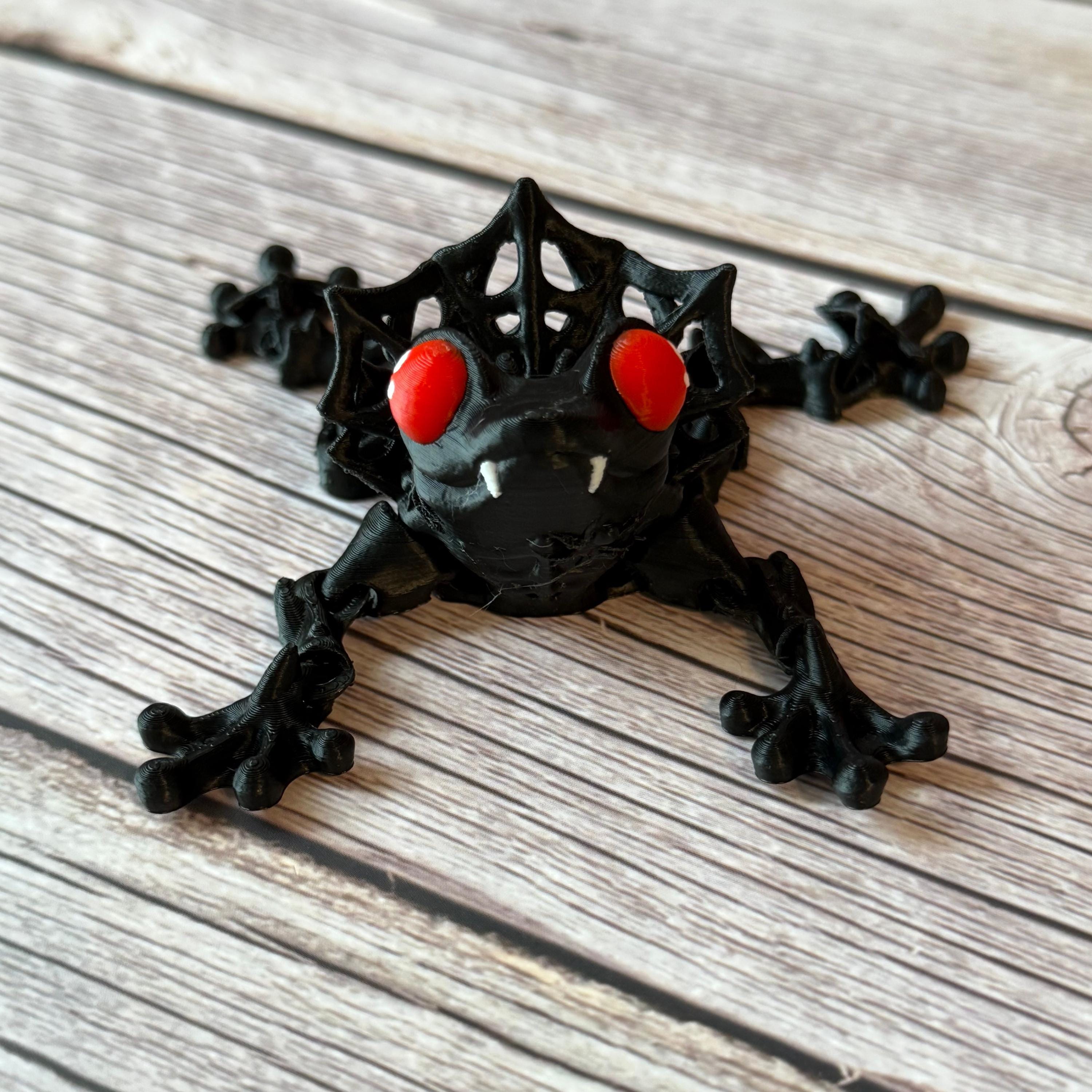 3D printed black and red frog with red eyes and vampire fangs, placed on a wooden surface