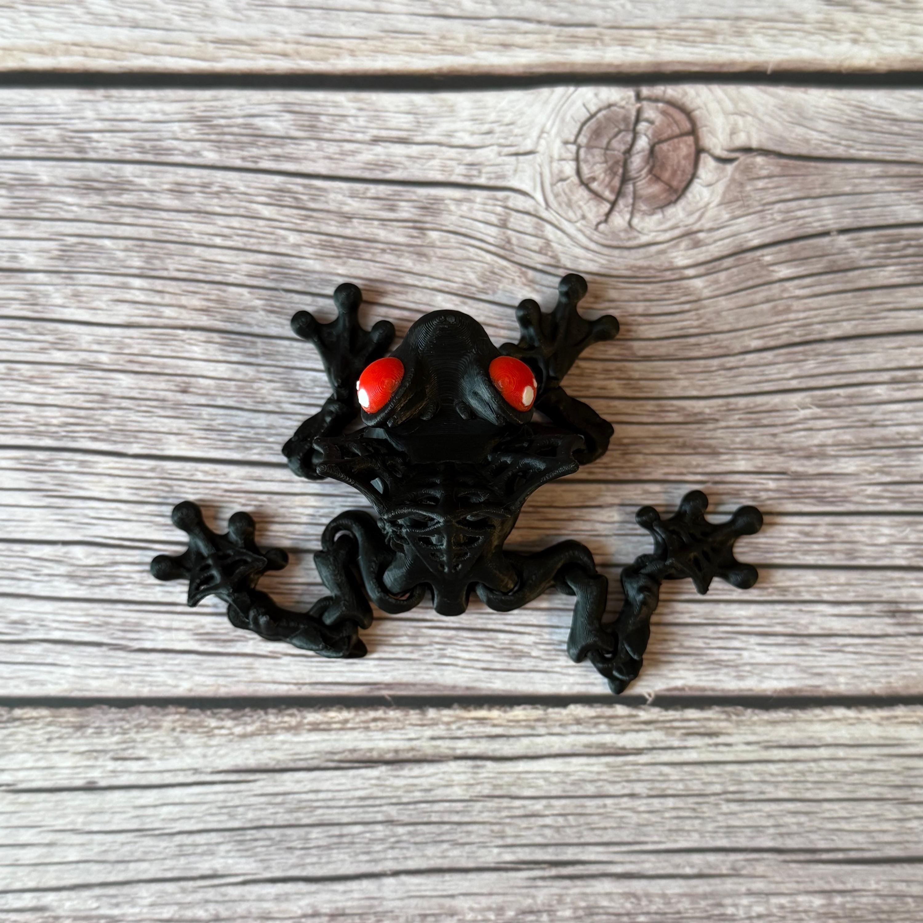 3D printed black and red frog with red eyes and vampire fangs, placed on a wooden surface