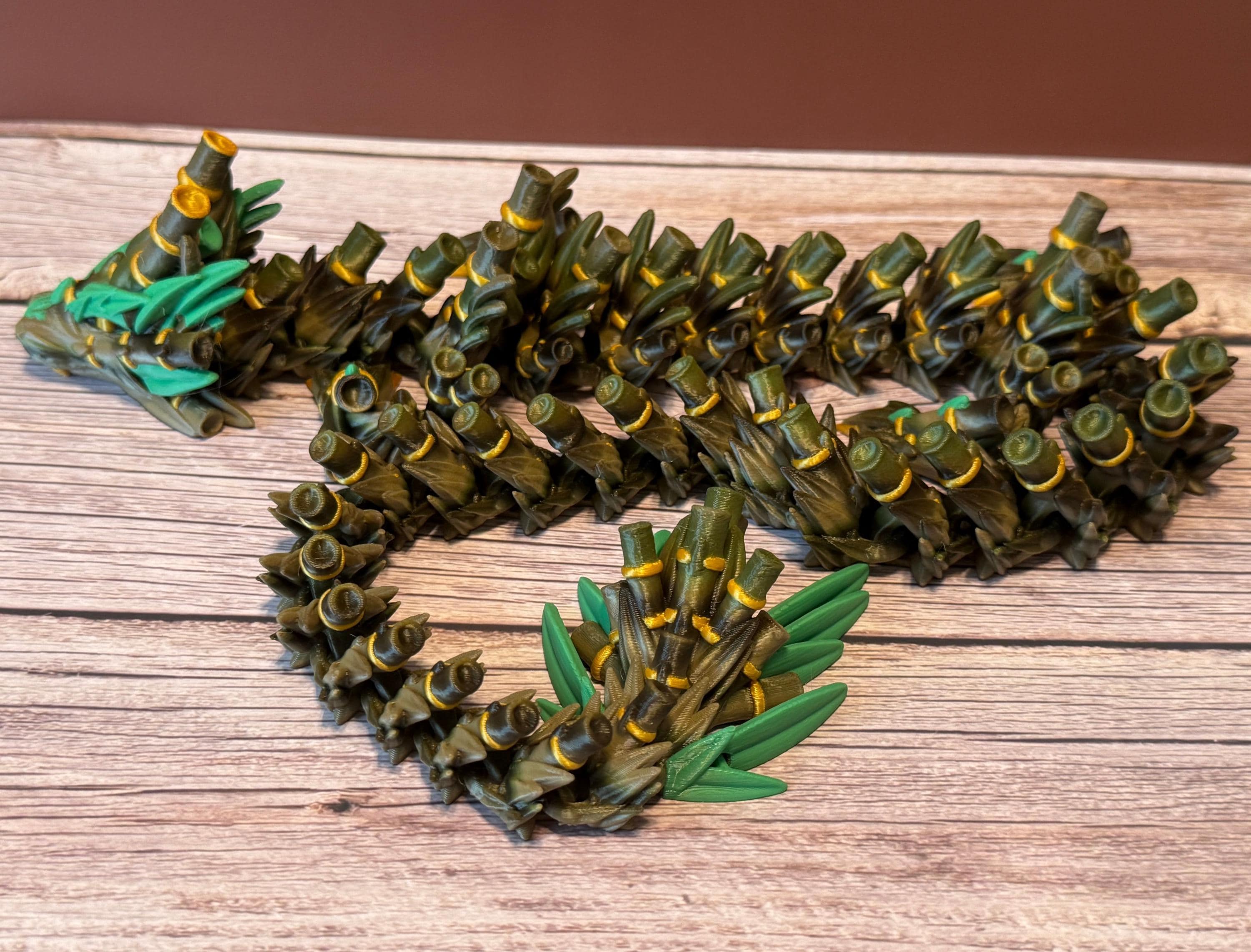 A green, brown, and gold colored 3D printed dragon figure with bamboo-like features, placed on a wooden surface