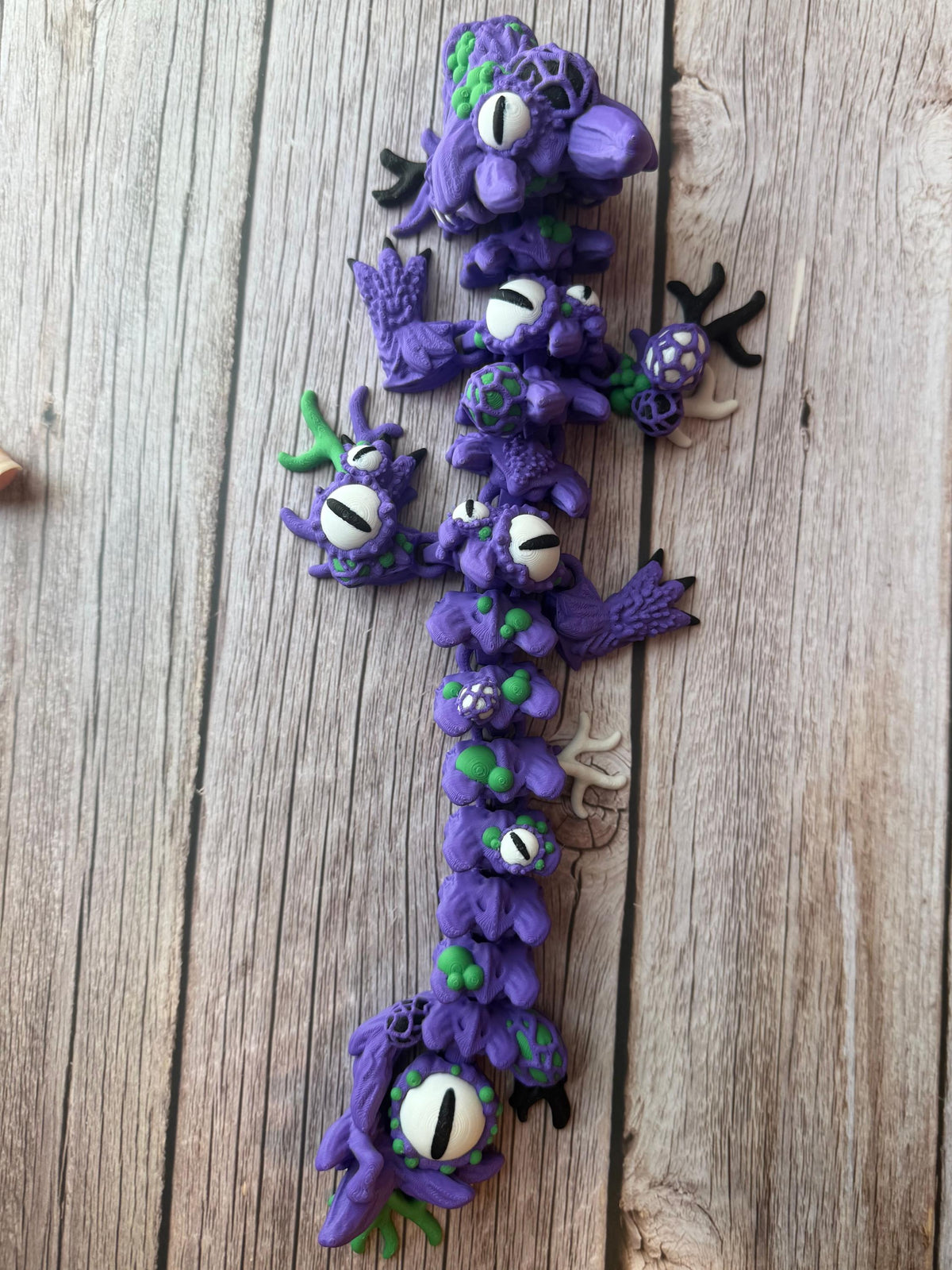 Purple and green 3D printed dragon with multiple eyes and growth-like features all over its body, placed on a wooden surface