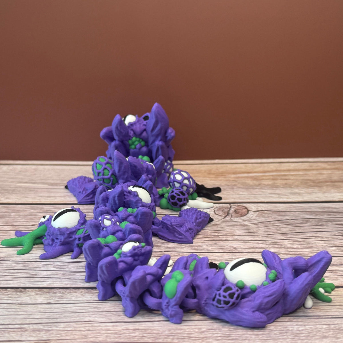 Purple and green 3D printed dragon with multiple eyes and growth-like features all over its body, placed on a wooden surface