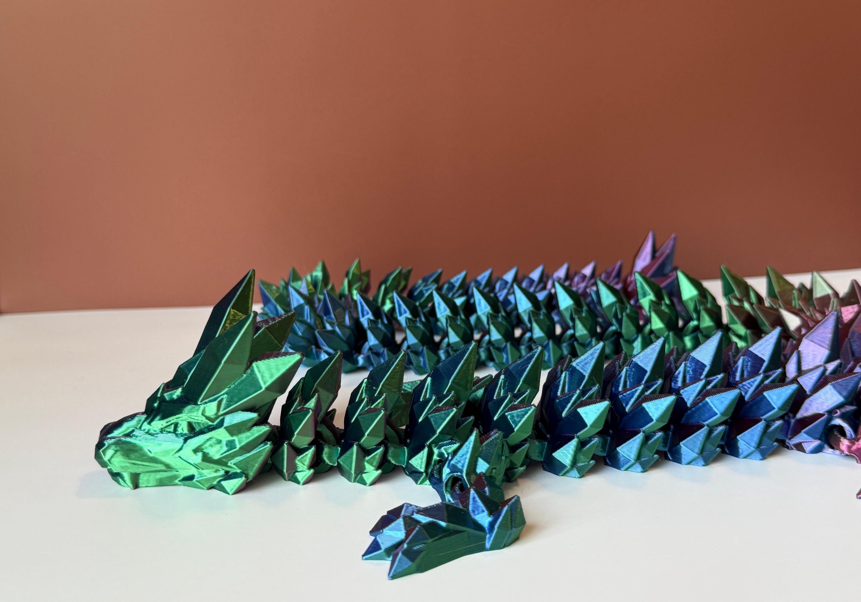 Multicolored 3D printed dragon with crystal-shaped features across the entire body, placed on a white surface with a brown backdrop