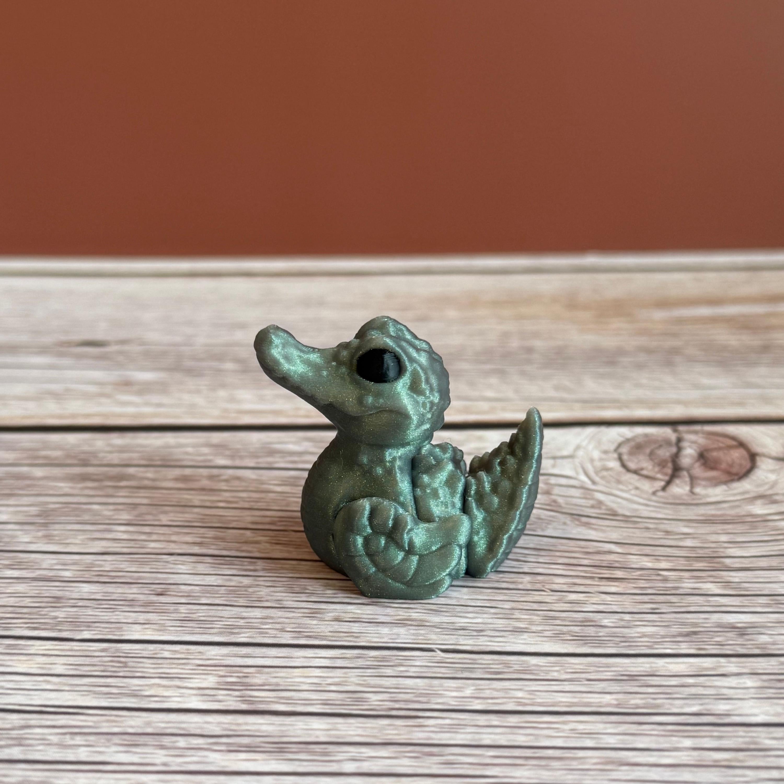 A small 3D printed figure depicting a hybrid animal resembling a duck and a crocodile, with a greenish tint, placed on a wooden surface.