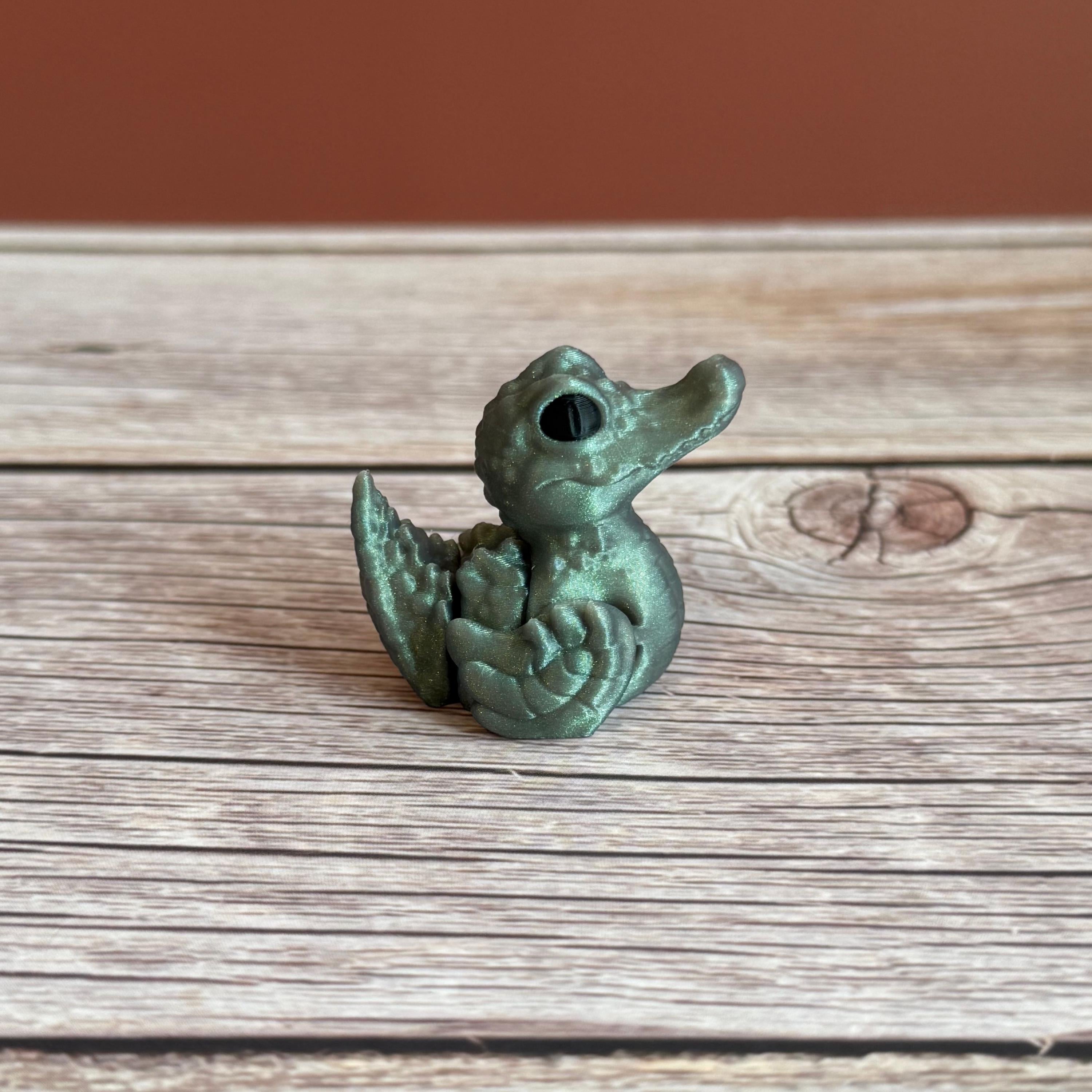 A small 3D printed figure depicting a hybrid animal resembling a duck and a crocodile, with a greenish tint, placed on a wooden surface.