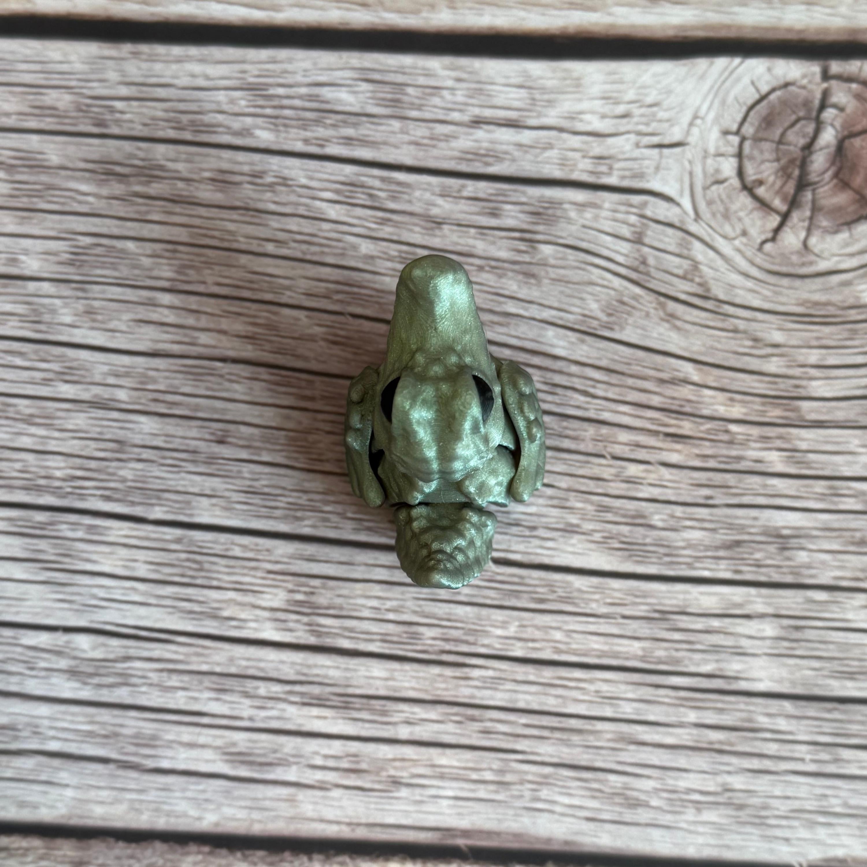 A small 3D printed figure depicting a hybrid animal resembling a duck and a crocodile, with a greenish tint, placed on a wooden surface.