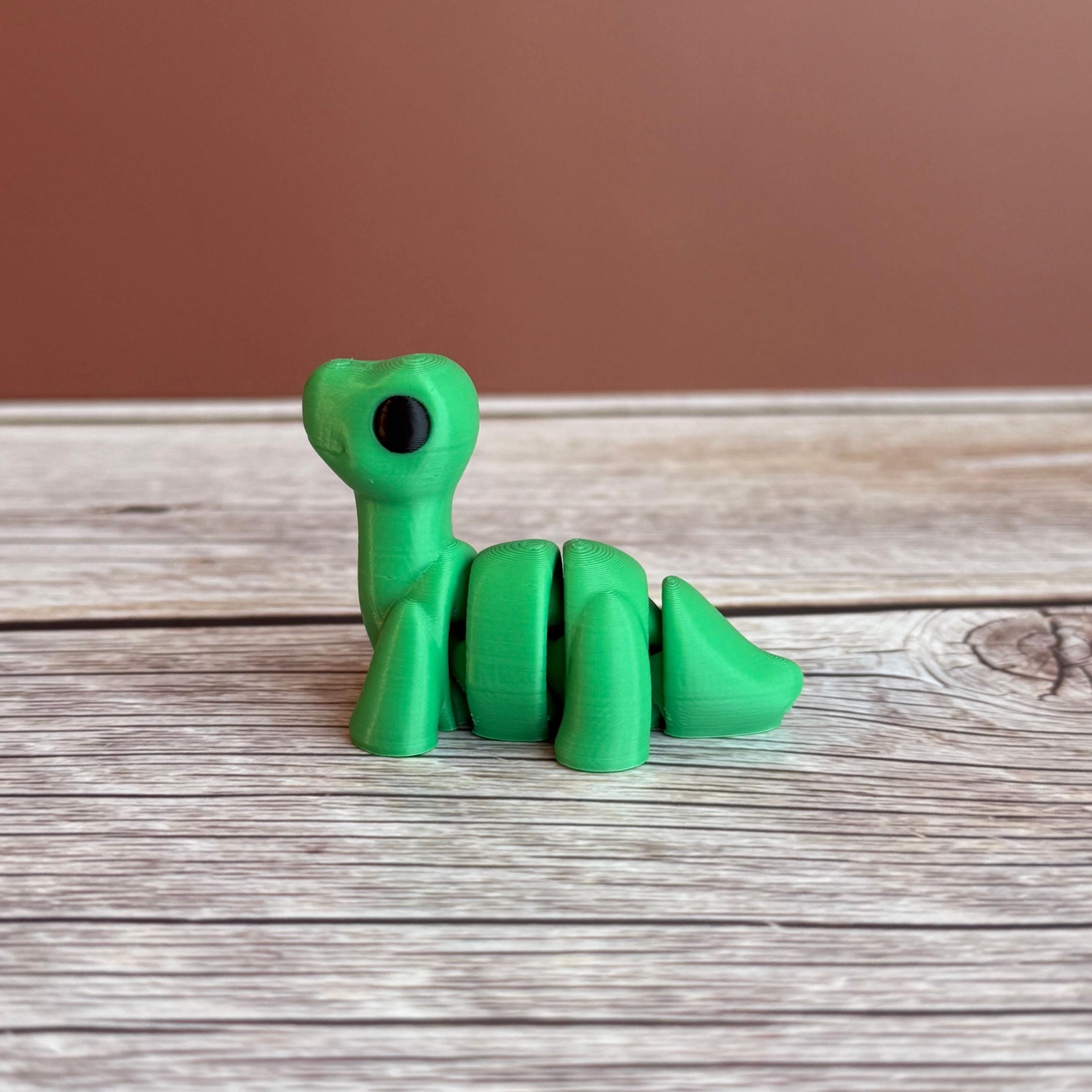 A small green 3D printed articulated Brontosaurus dinosaur toy, sitting on a wooden surface.