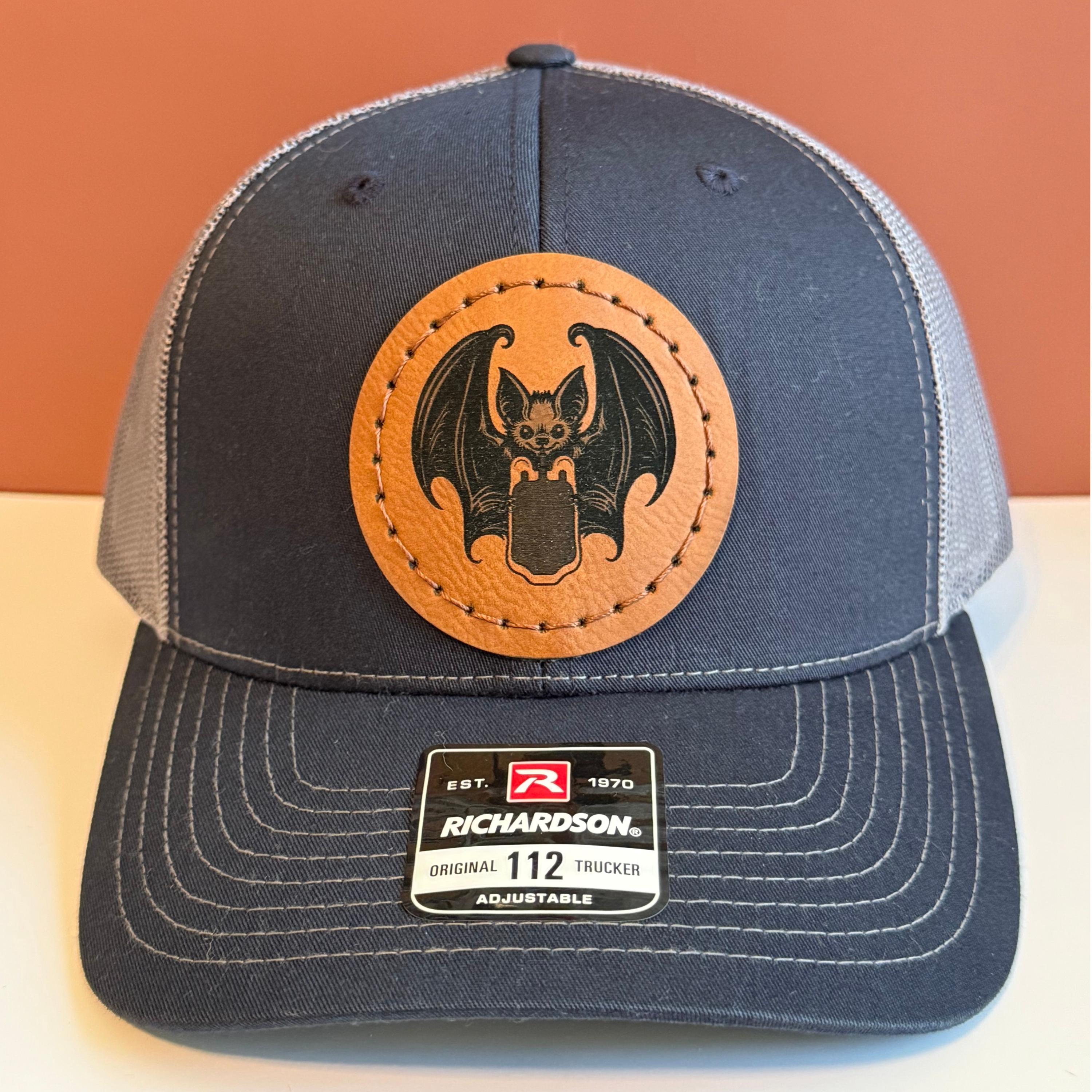 A blue and gray snapback hat with a hand-sewn leatherette patch featuring a vampire bat holding a blood bag, placed against a white and brown background