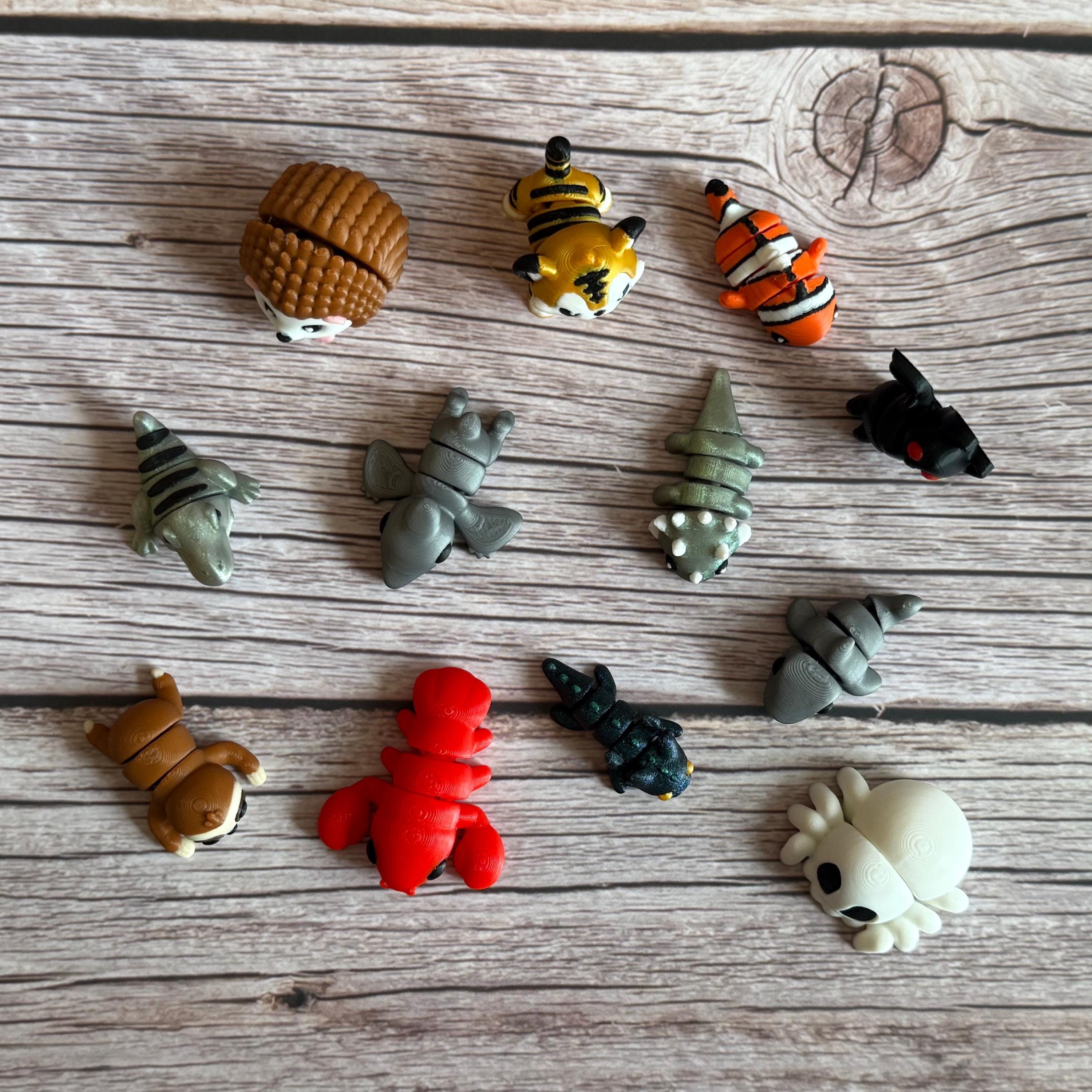 Collection of small animal figurines on a wooden surface. T-rex, hedgehog, sloth, pterodactyl, tiger, lobster, dragon, triceratops, clown fish, bat, shark, and spider-shaped skeleton are featured. 