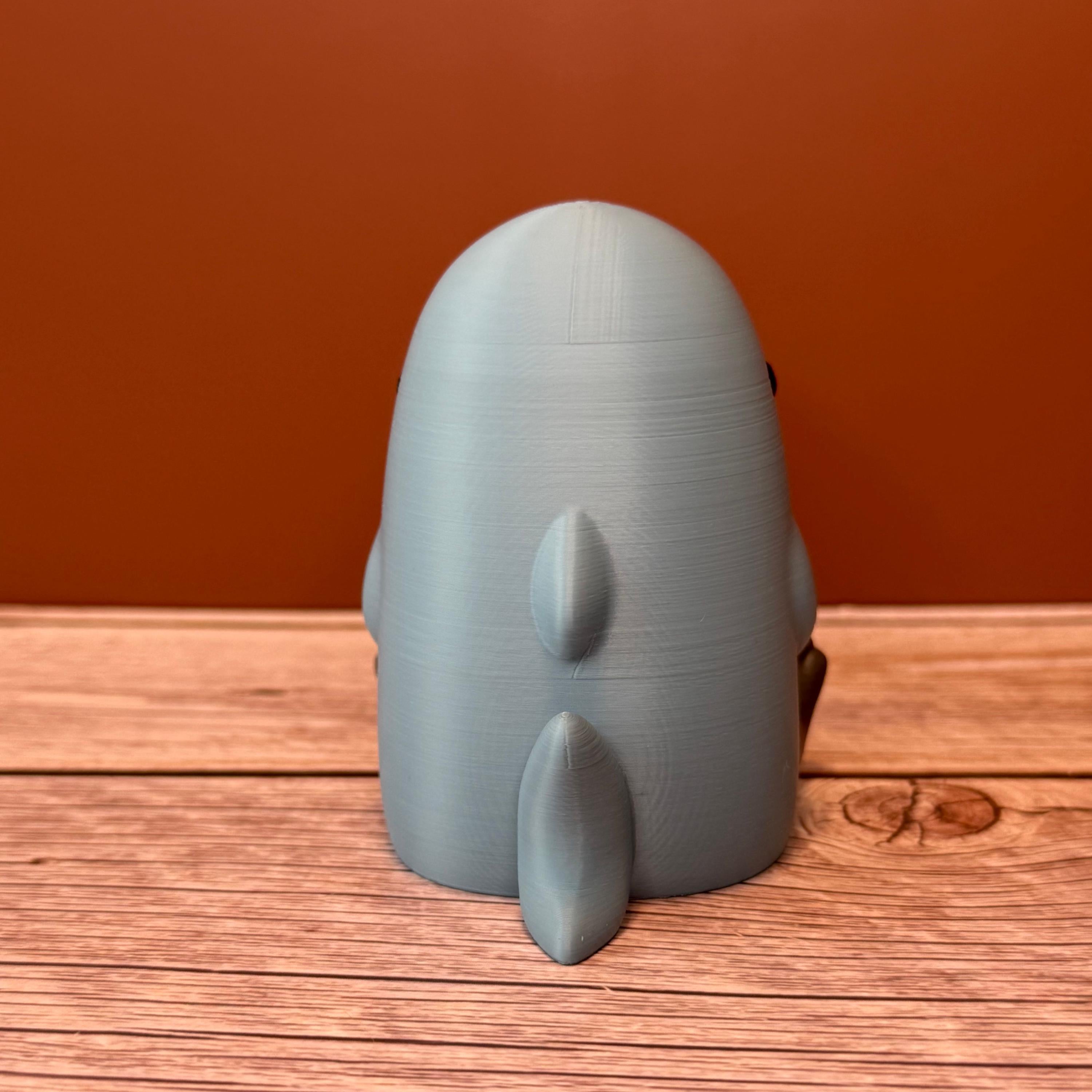 Backside of a 3D printed figurine of a cat wearing a shark costume in blue-gray color, placed on a wooden surface