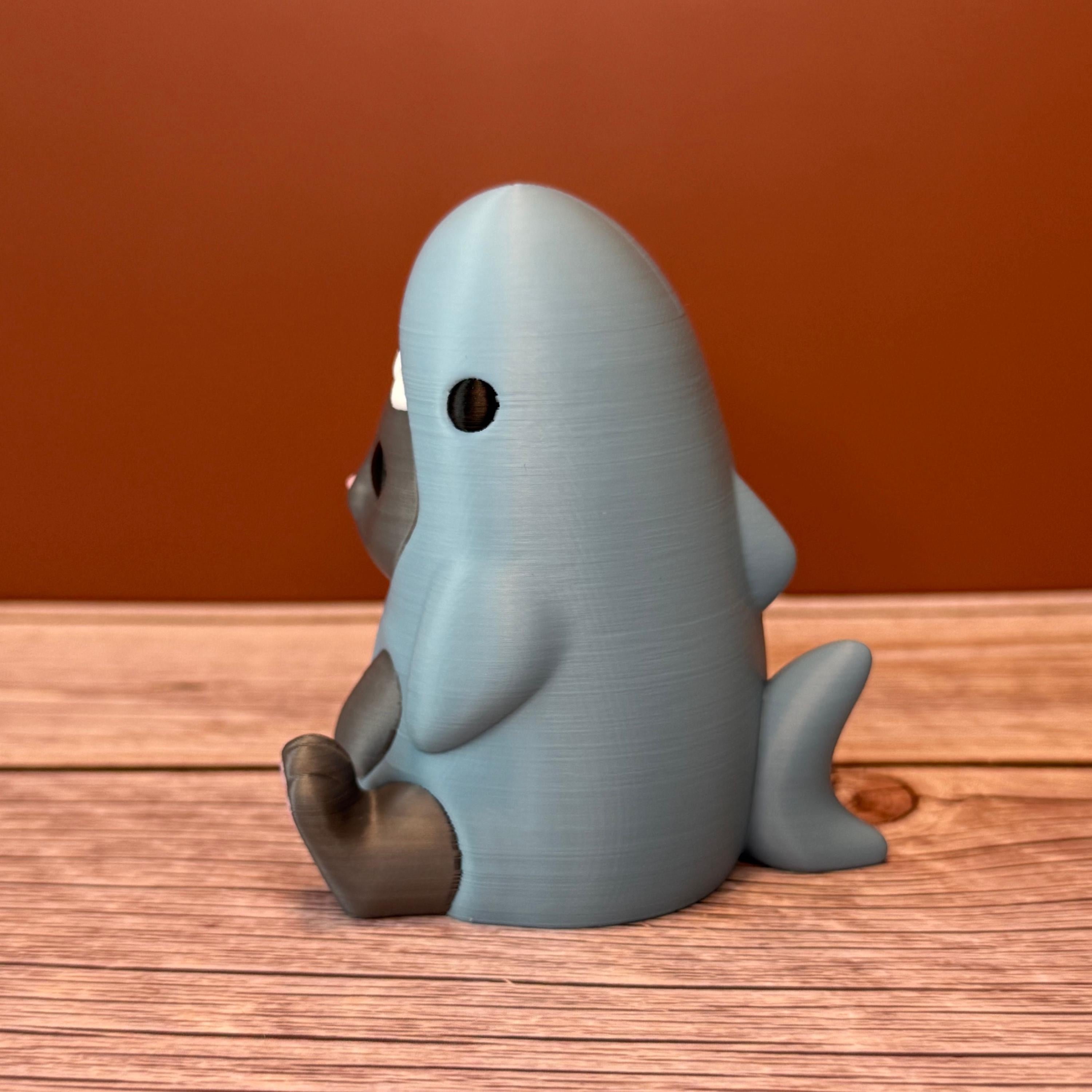 3D printed figurine of a gray cat wearing a shark costume in blue-gray color, placed on a wooden surface