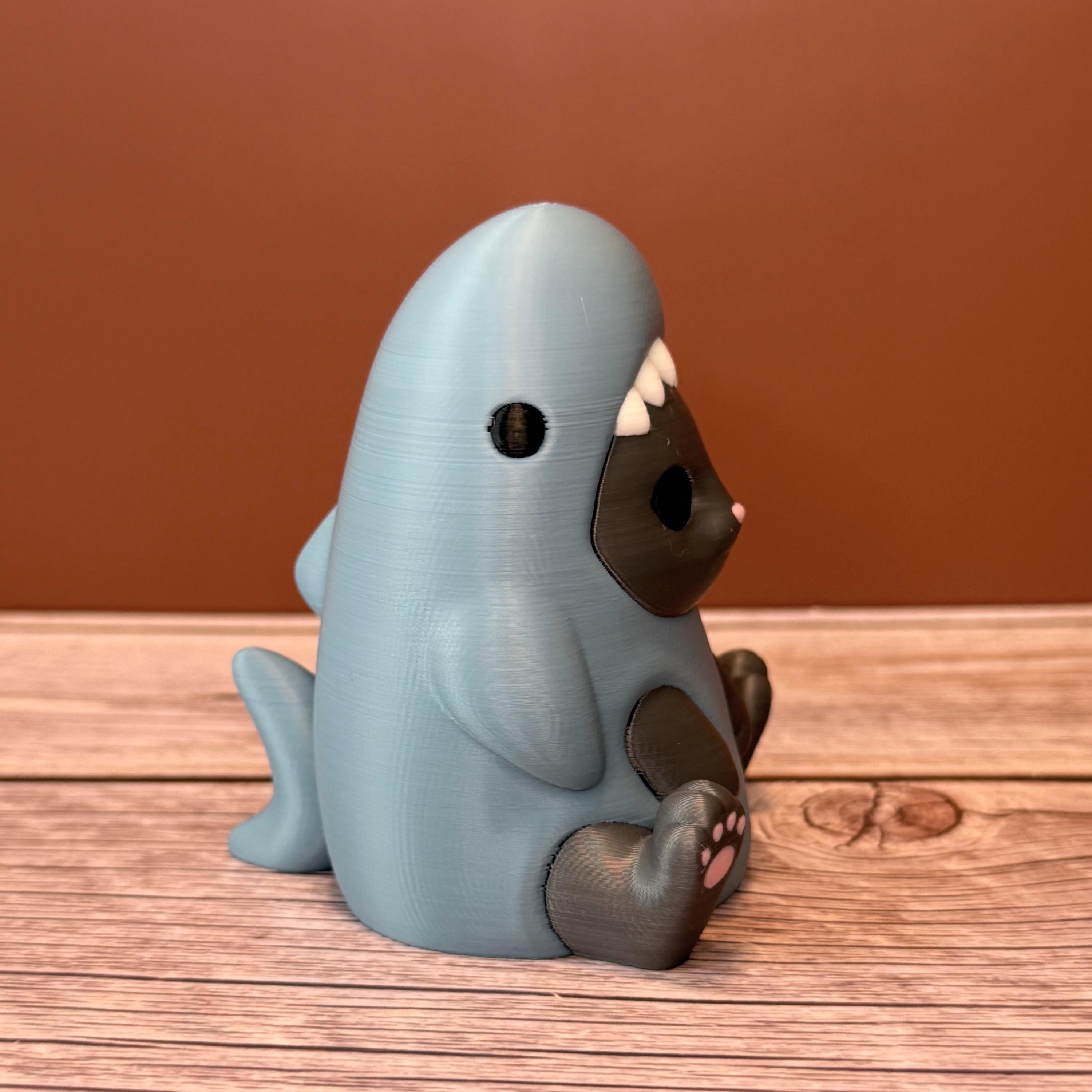 3D printed figurine of a gray cat wearing a shark costume in blue-gray color, placed on a wooden surface