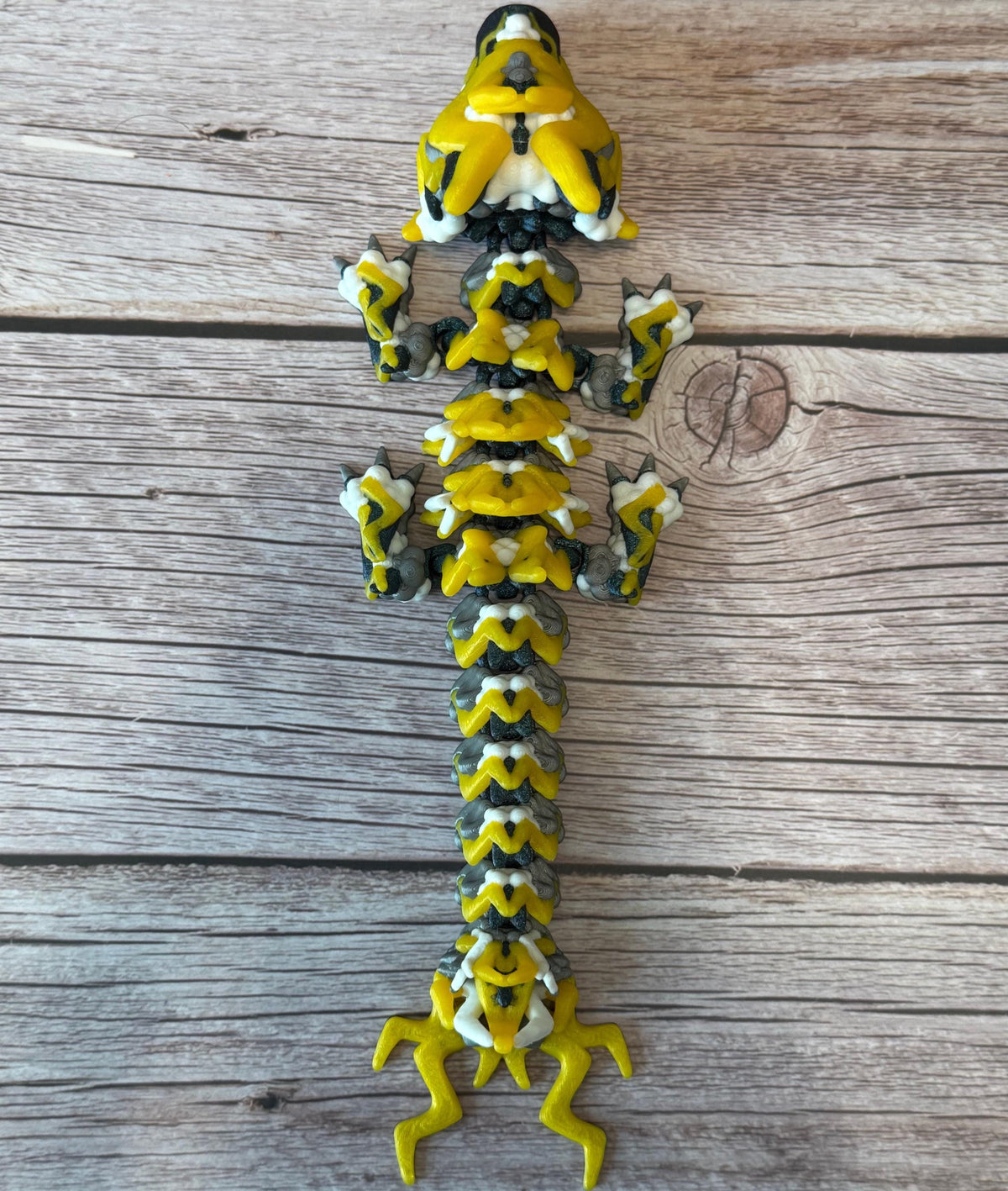 A 3D printed, articulated toy dragon figurine with yellow and black coloring, featuring lightning bolt details and storm cloud patterns, placed on a wooden surface