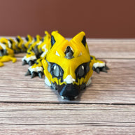 A 3D printed, articulated toy dragon figurine with yellow and black coloring, featuring lightning bolt details and storm cloud patterns, placed on a wooden surface