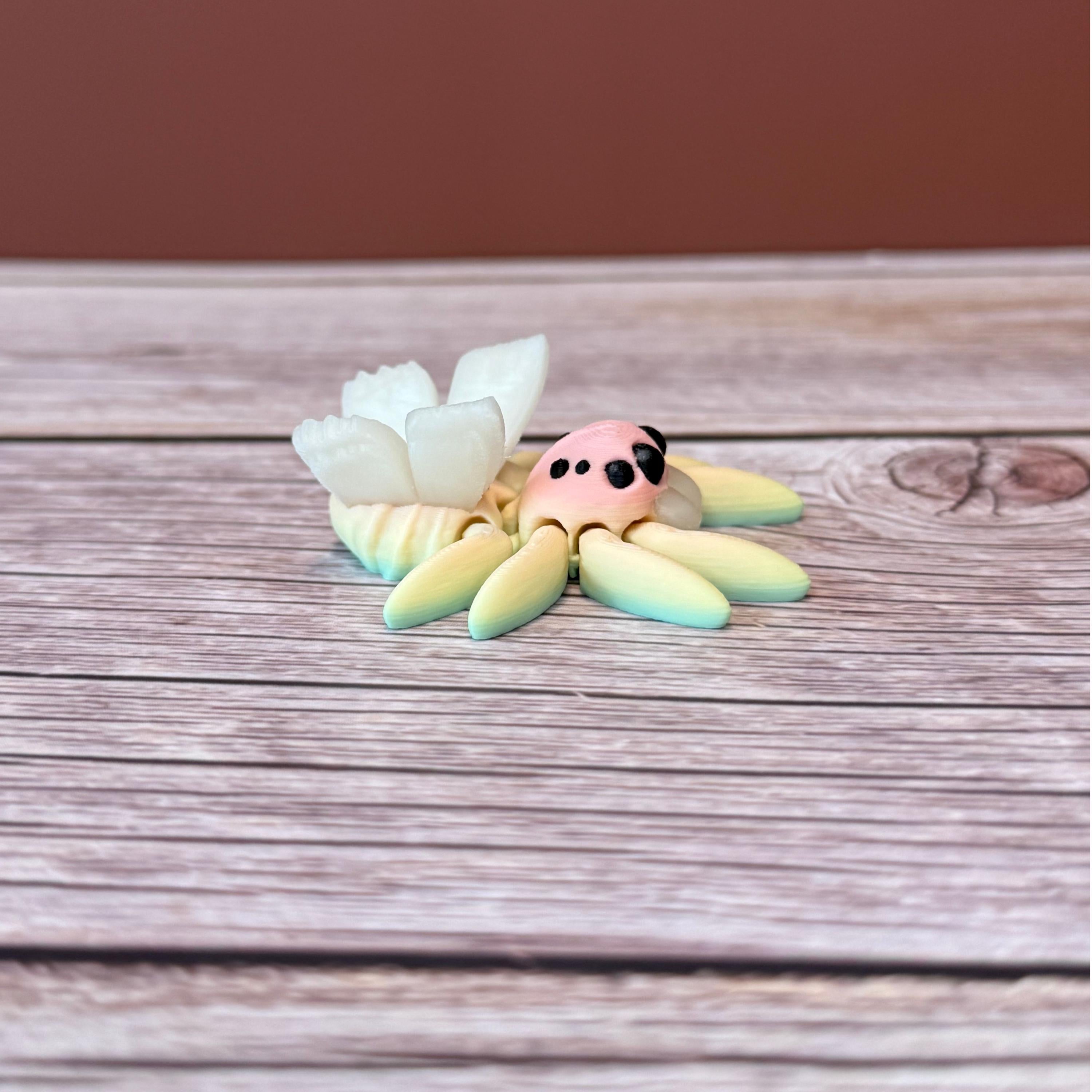 3D printed small spider with fairy design featuring rainbow coloring and white wings, placed on a wooden surface