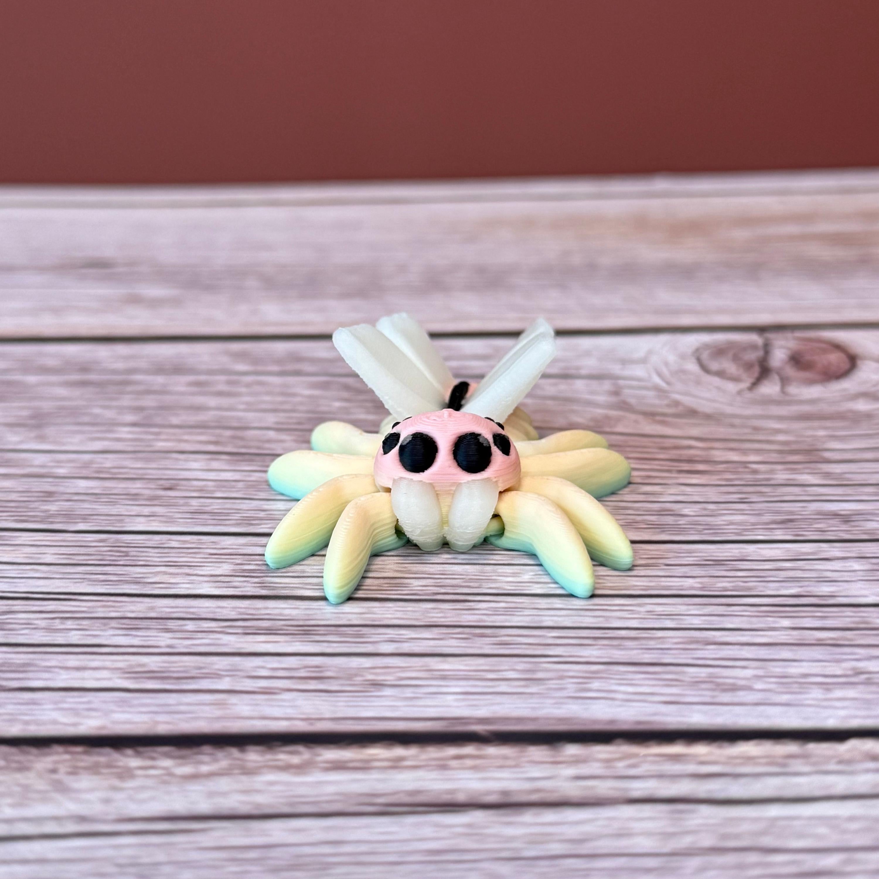 3D printed small spider with fairy design featuring rainbow coloring and white wings, placed on a wooden surface