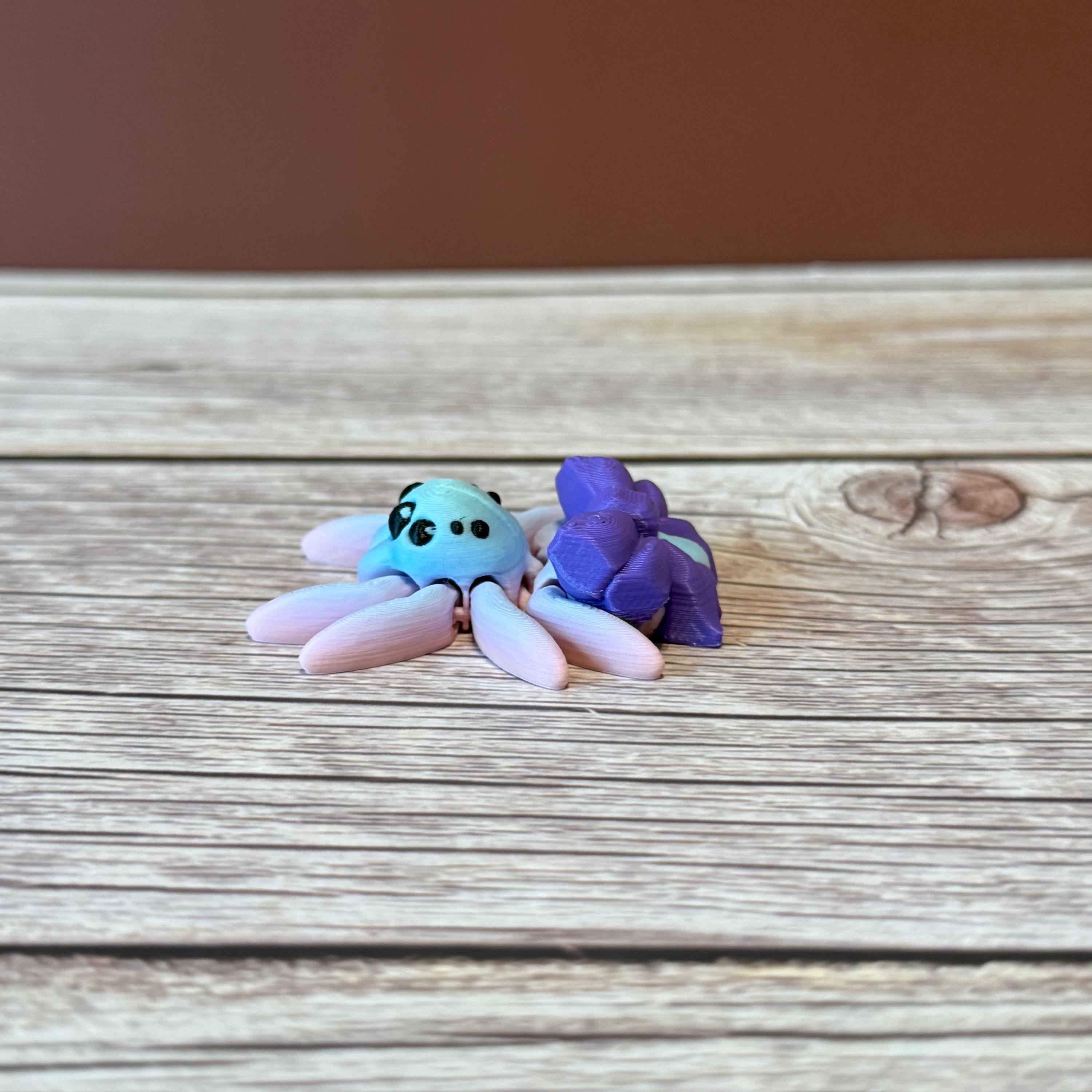 A 3D printed toy figurine of an articulated spider with a giant purple bow and multicolored details on a wooden surface.