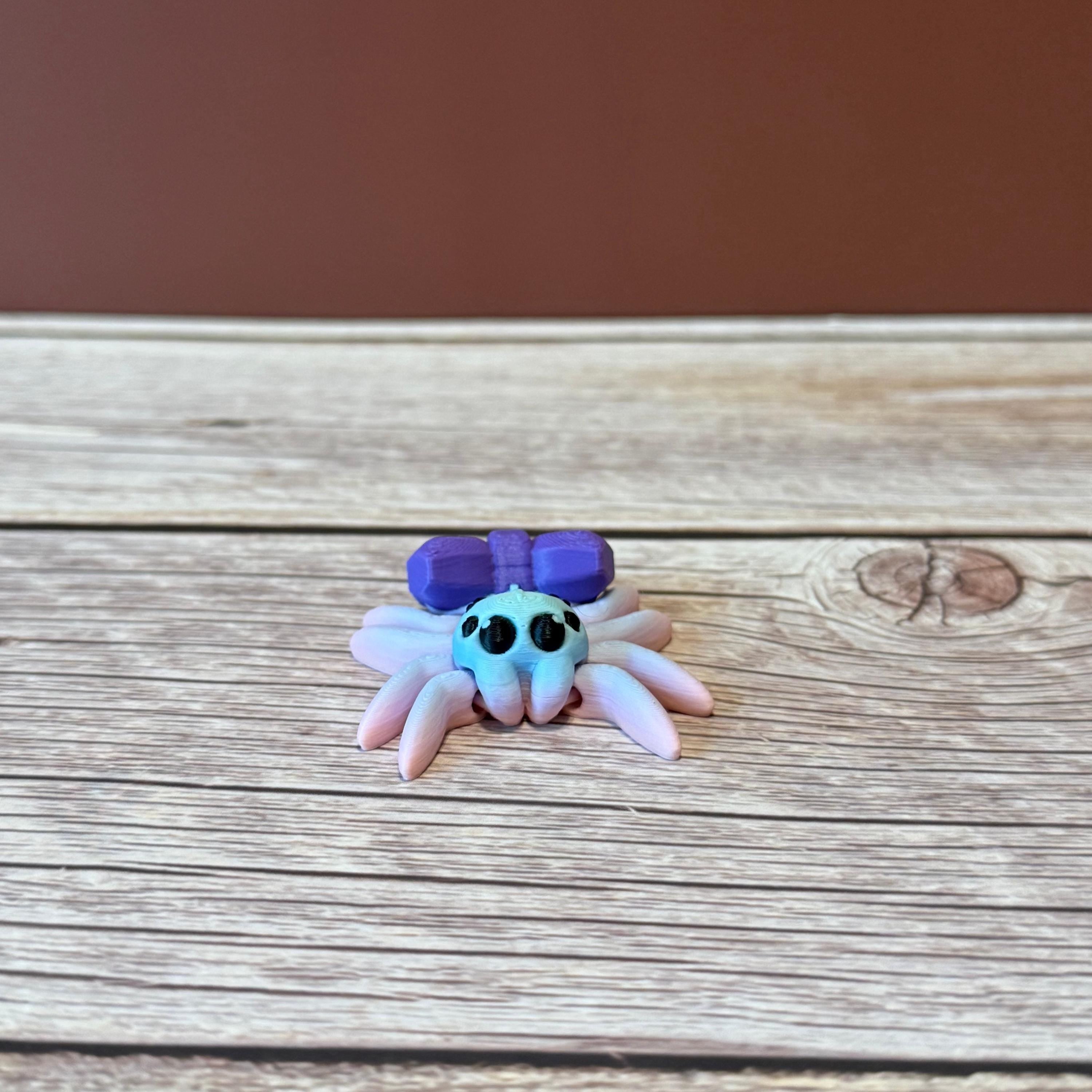 A 3D printed toy figurine of an articulated spider with a giant purple bow and multicolored details on a wooden surface.