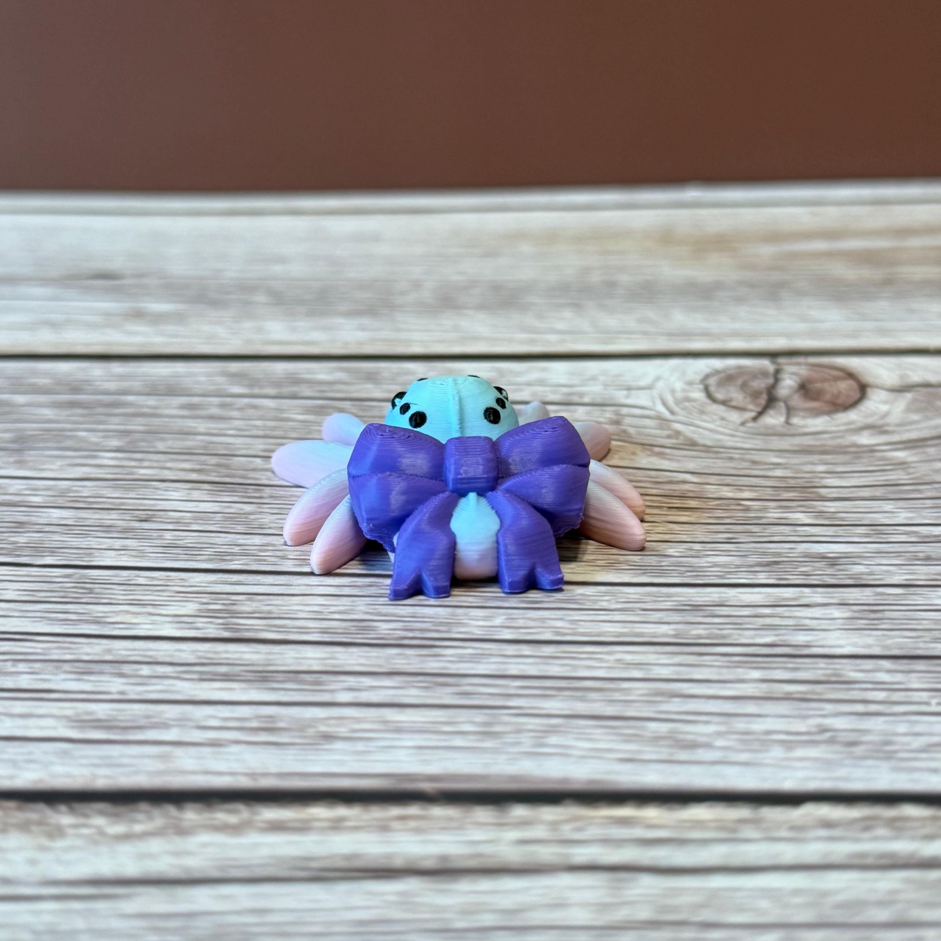 A 3D printed toy figurine of an articulated spider with a giant purple bow and multicolored details on a wooden surface.