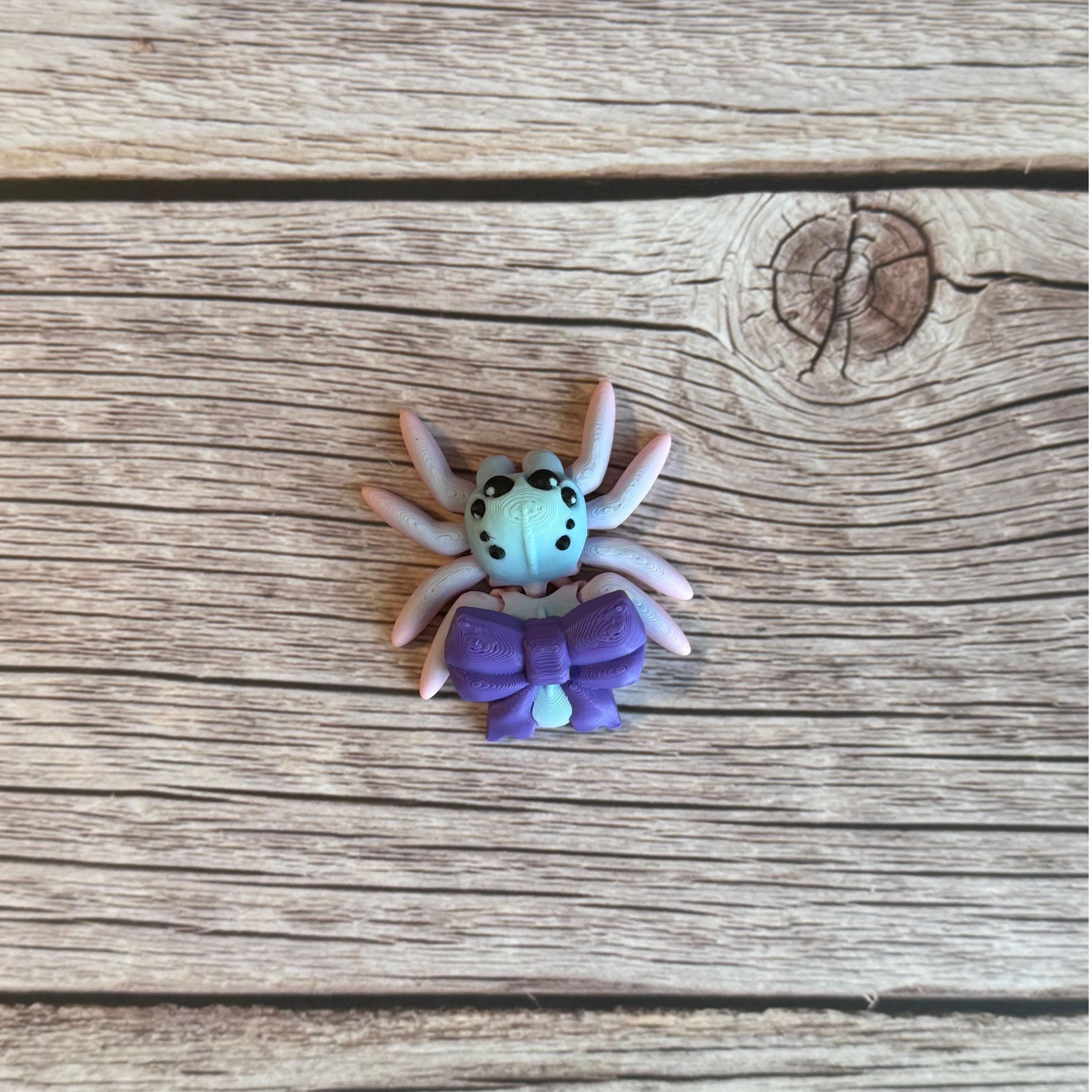 A 3D printed toy figurine of an articulated spider with a giant purple bow and multicolored details on a wooden surface.