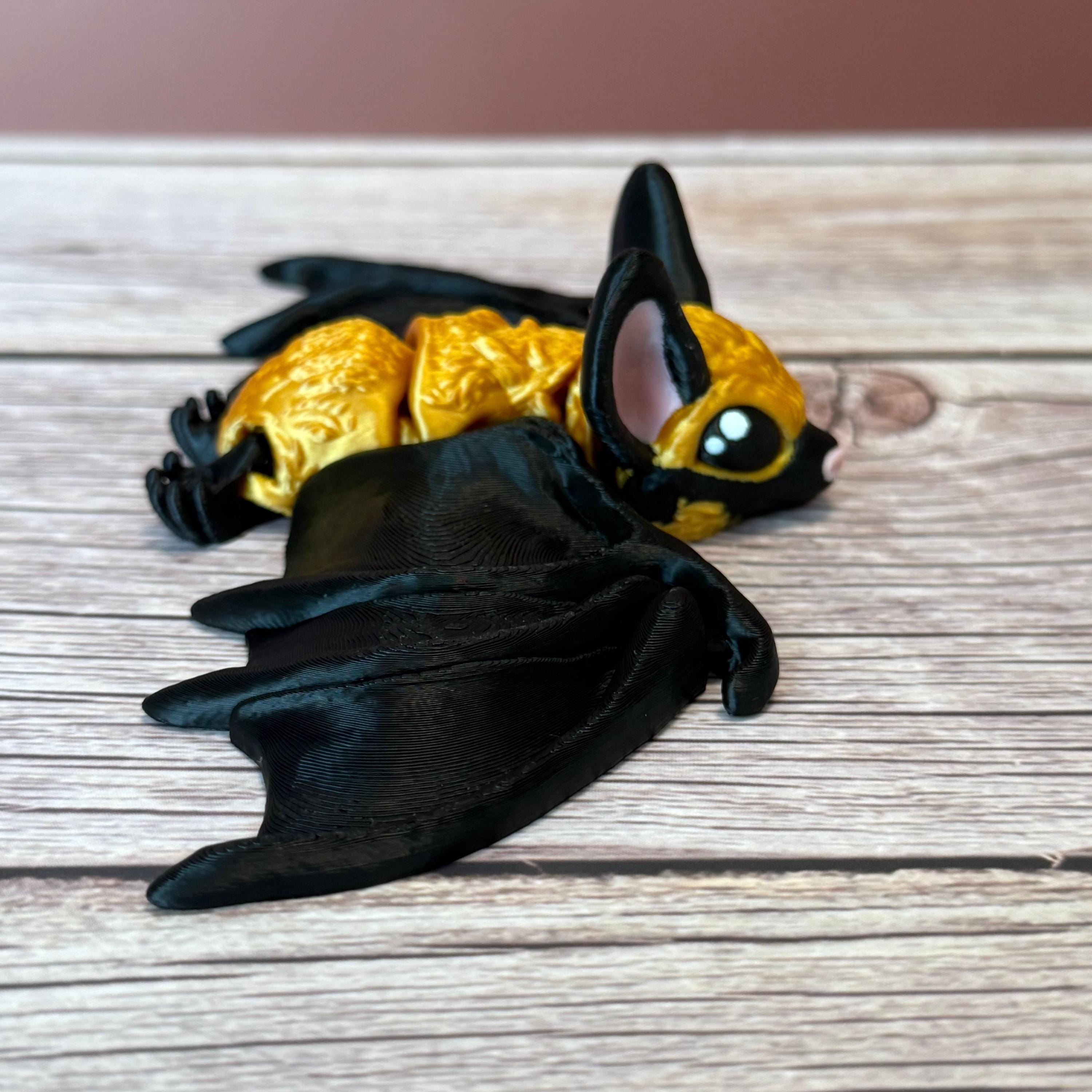 3D printed gold and black articulated bat with tall pink ears and pink nose, placed on a wooden surface