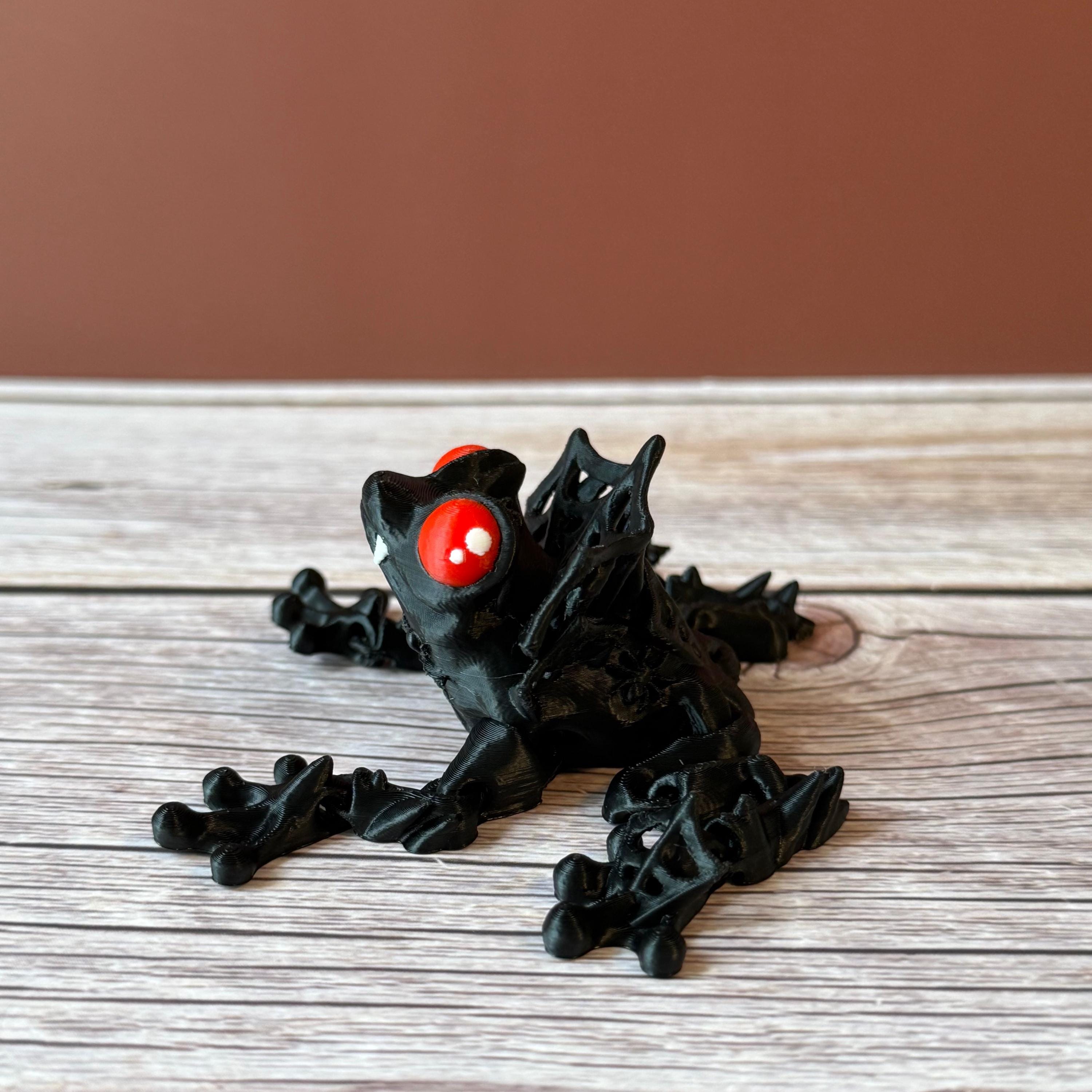 3D printed black and red frog with red eyes and vampire fangs, placed on a wooden surface