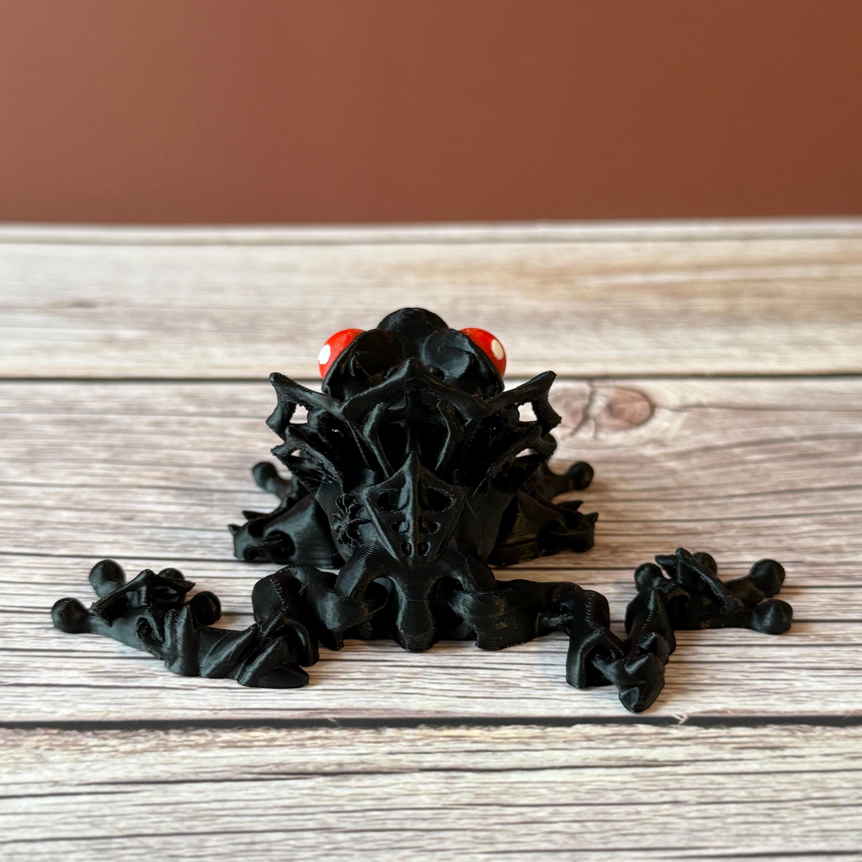 3D printed black and red frog with red eyes and spider webs on its back, placed on a wooden surface