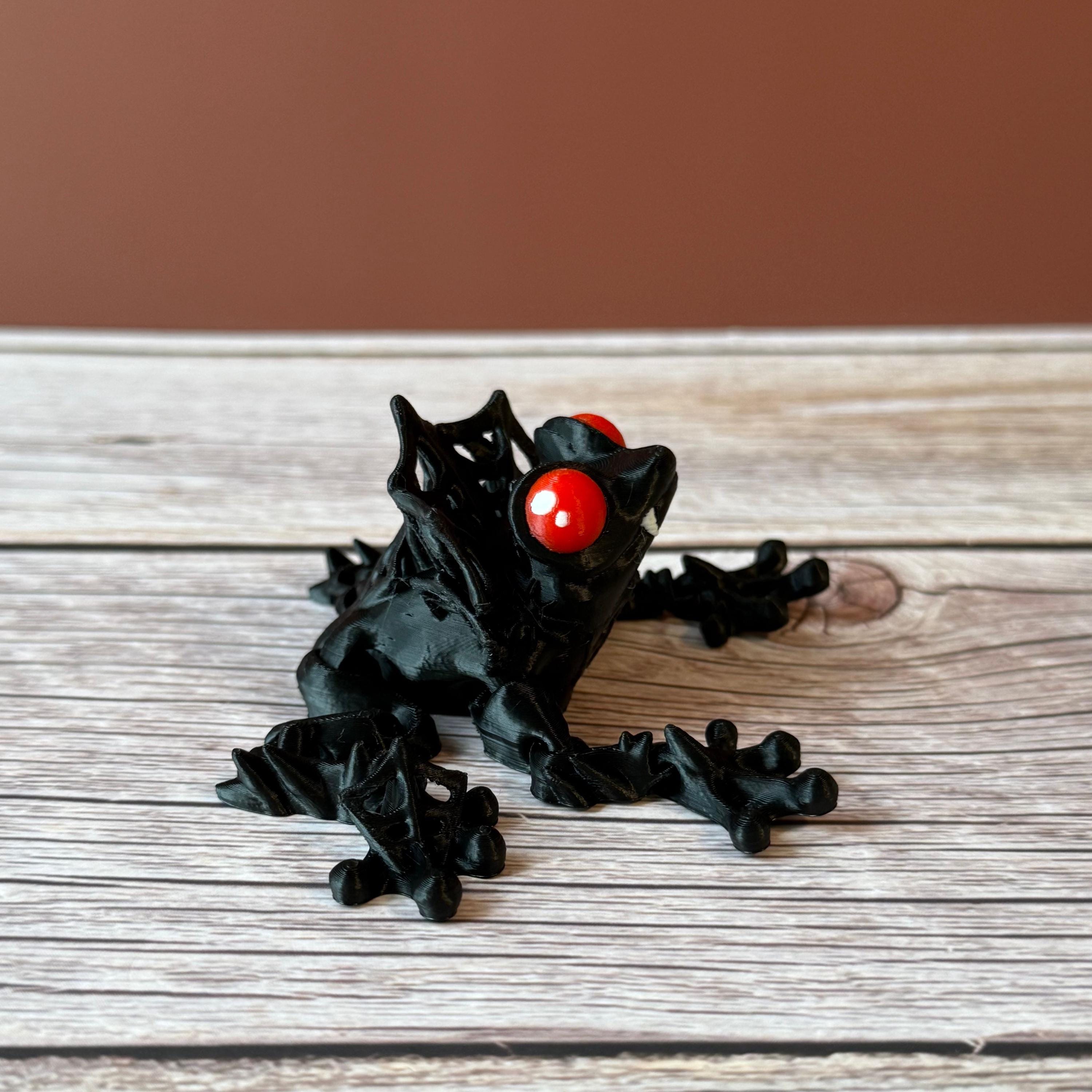 3D printed black and red frog with red eyes and vampire fangs, placed on a wooden surface