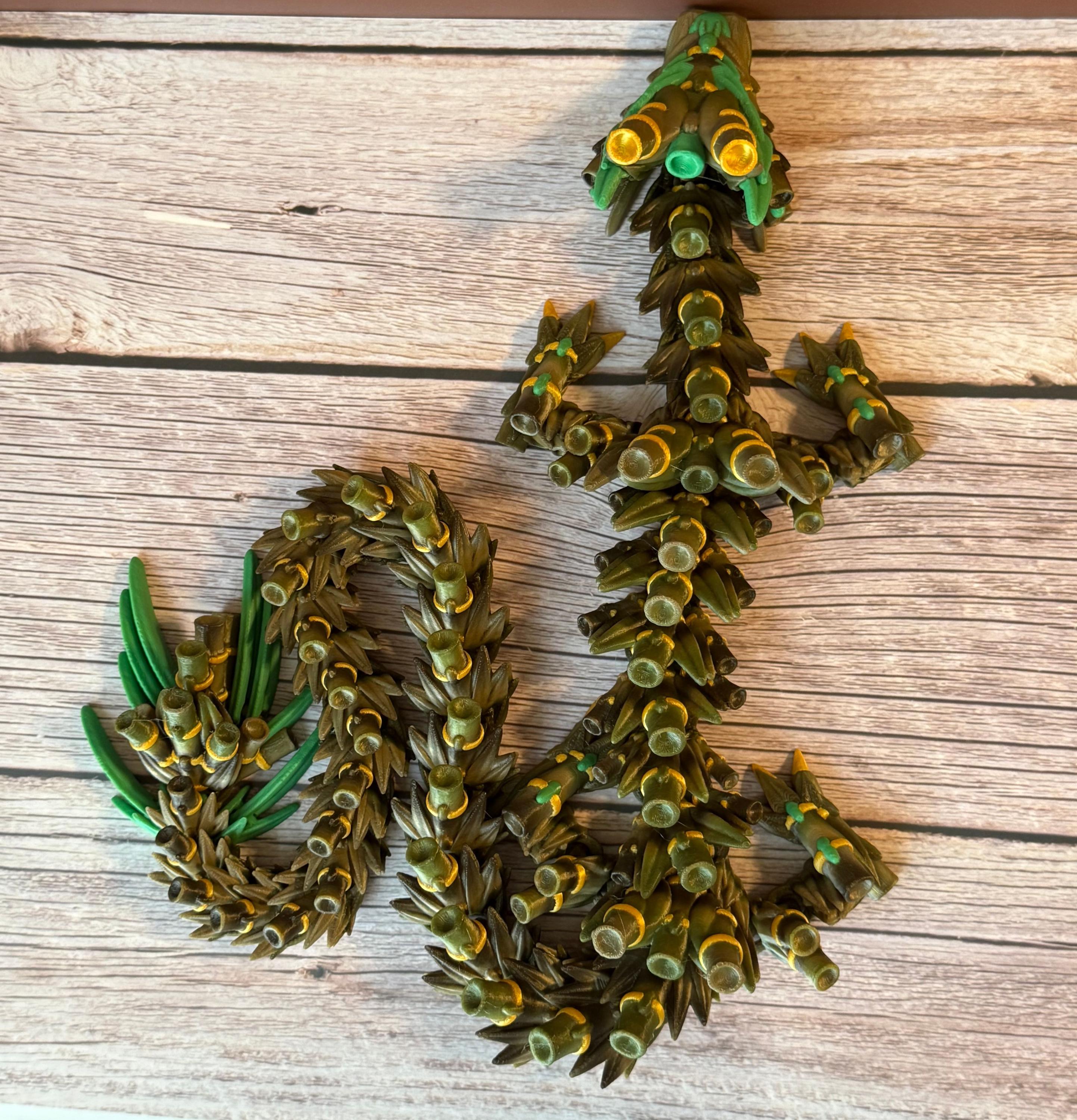 A green, brown, and gold colored 3D printed dragon figure with bamboo-like features, placed on a wooden surface