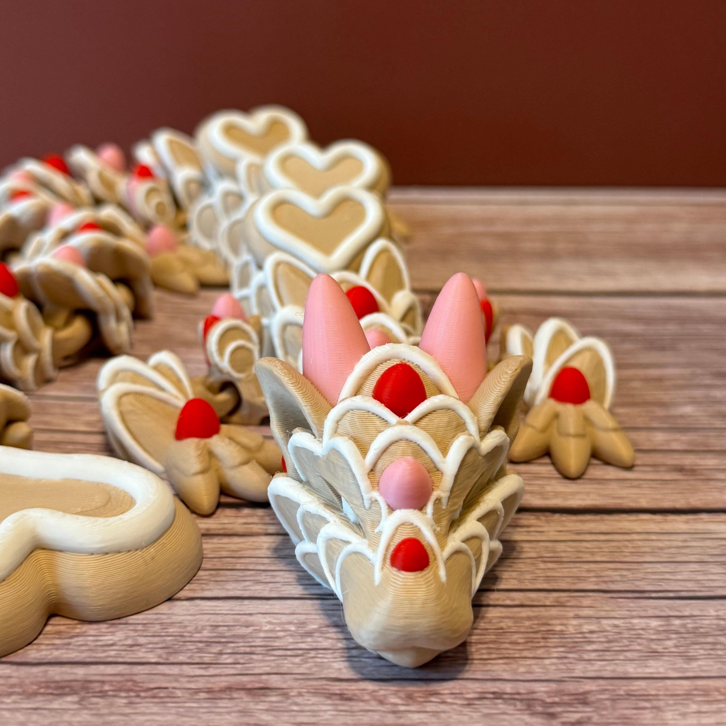 A beige colored 3D printed dragon figure with red, pink, and white heart-shaped cookie-like features, placed on a wooden surface