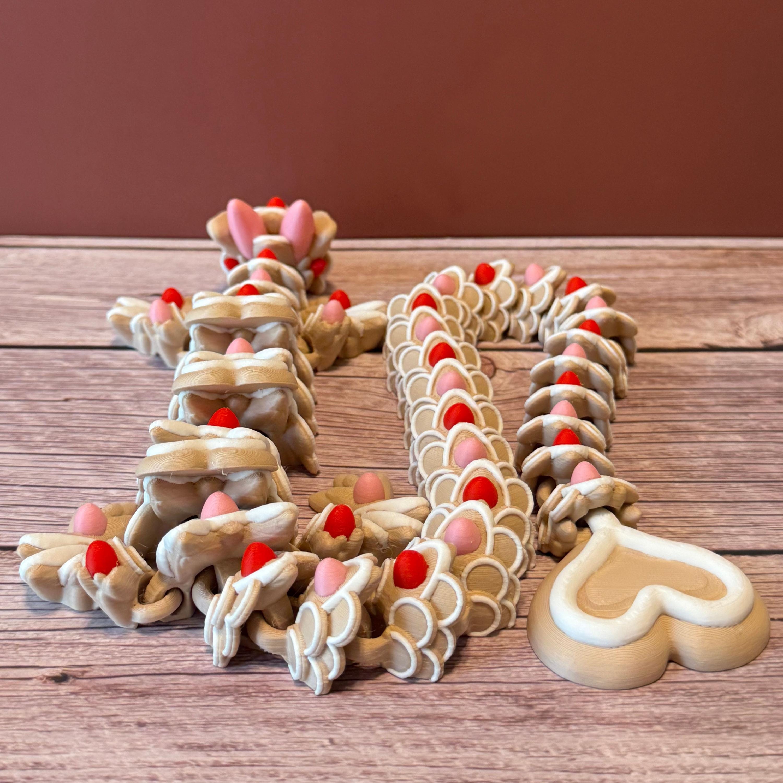A beige colored 3D printed dragon figure with red, pink, and white heart-shaped cookie-like features, placed on a wooden surface