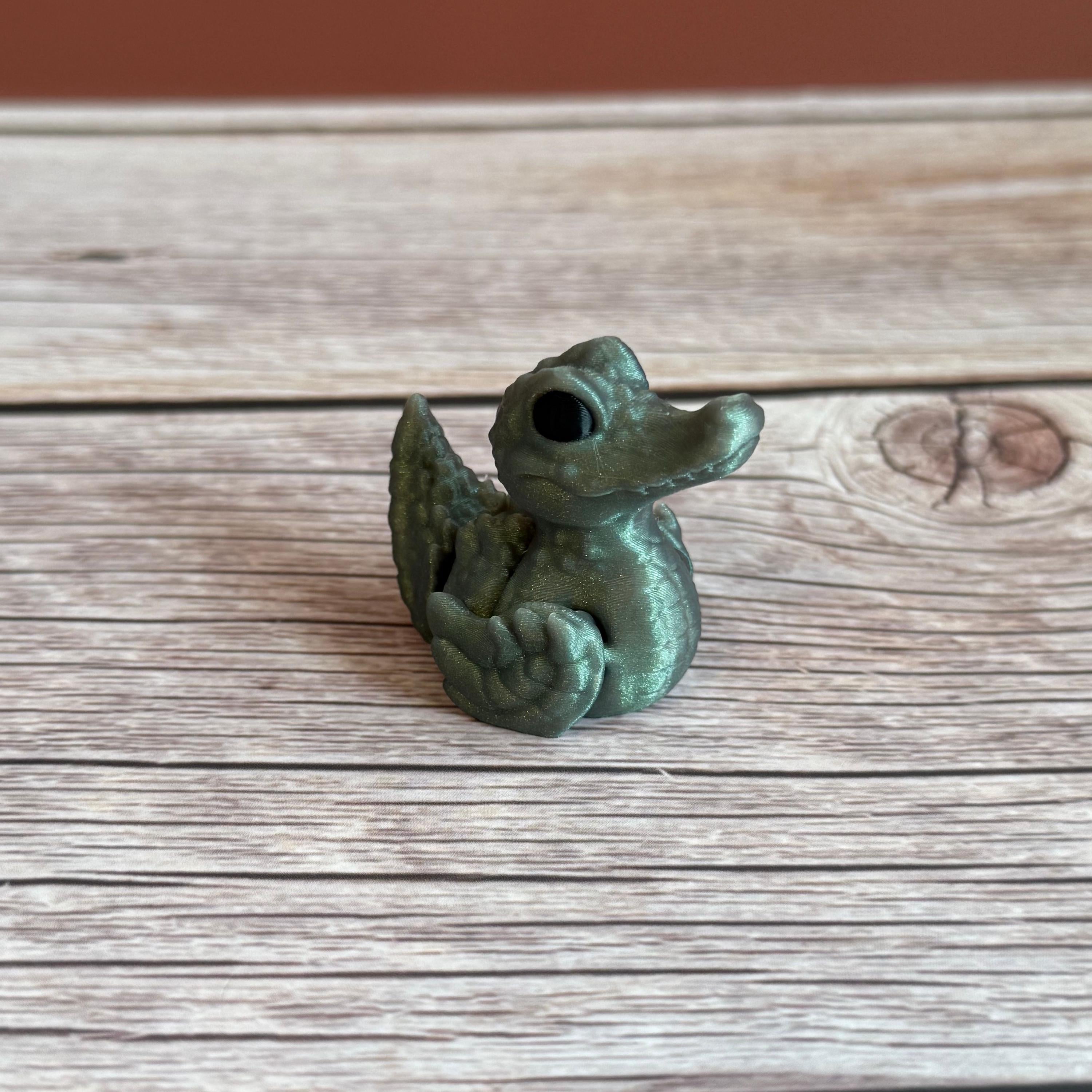 A small 3D printed figure depicting a hybrid animal resembling a duck and a crocodile, with a greenish tint, placed on a wooden surface.
