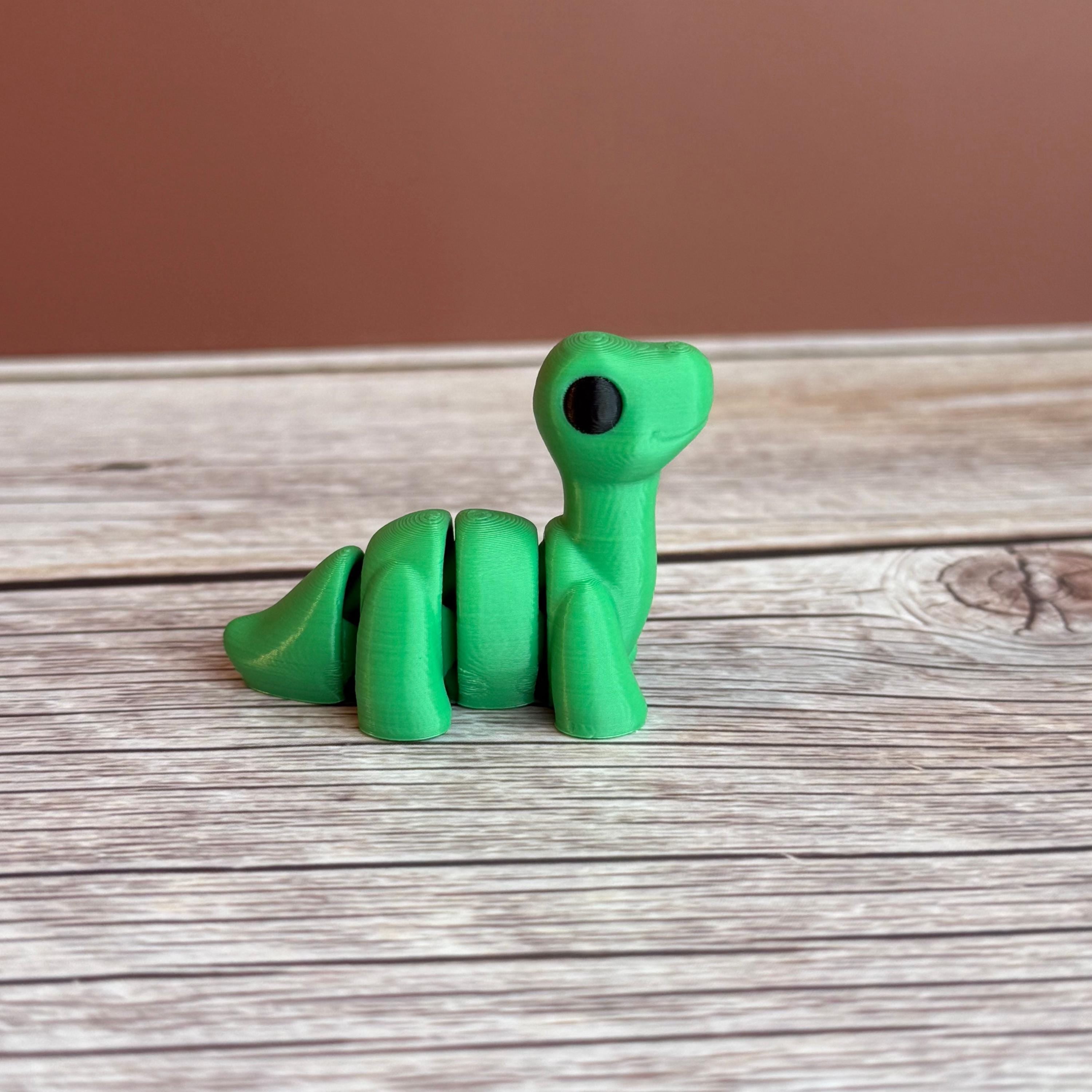 A small green 3D printed articulated Brontosaurus dinosaur toy, sitting on a wooden surface.