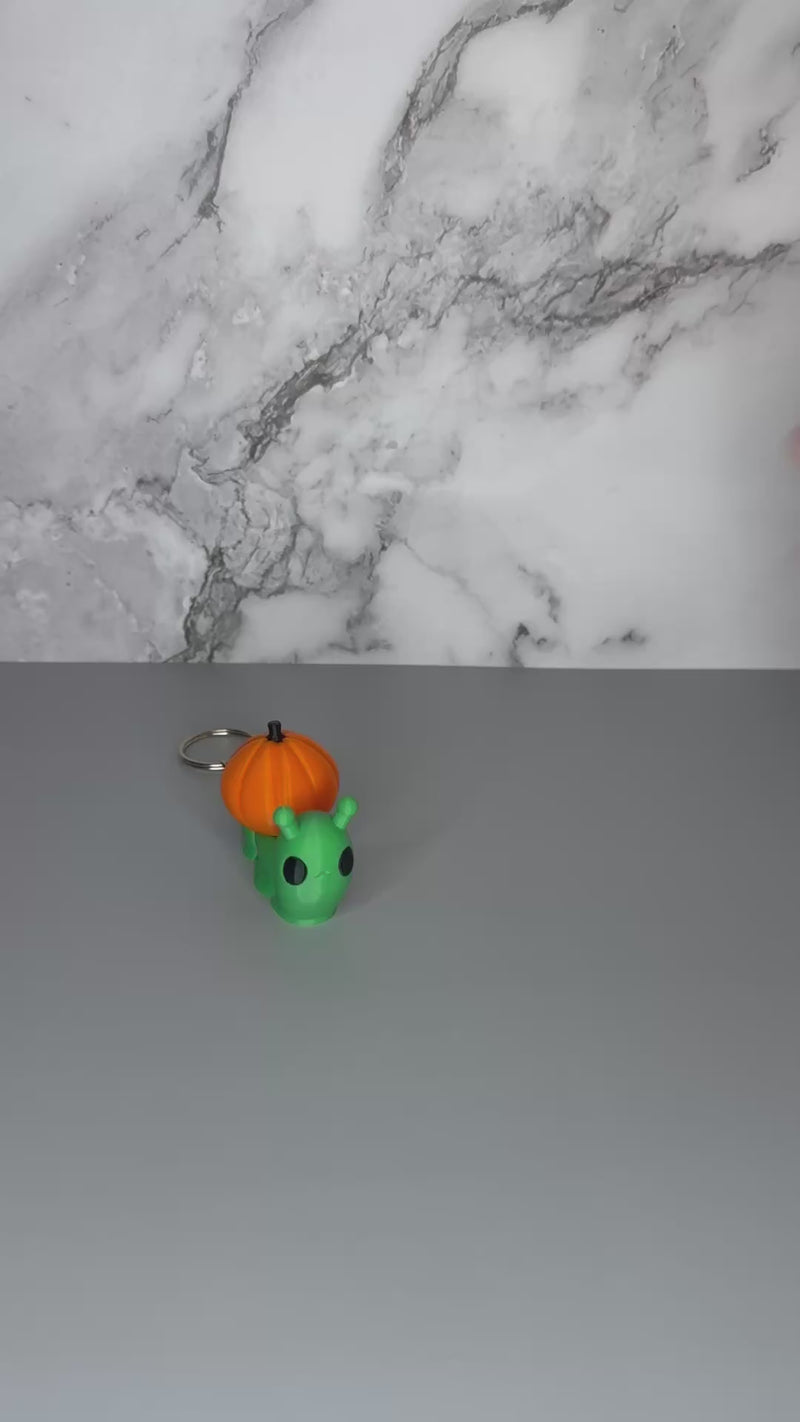 Green snail-shaped keychain with orange jack-o-lantern shell on a gray marble background being shaken to demonstrate articulation.