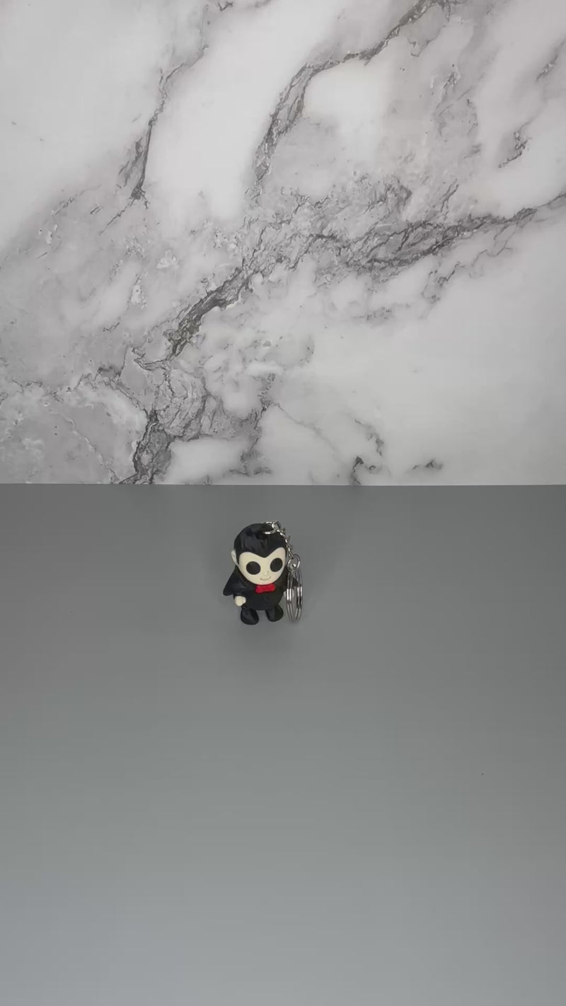 Keychain of a dracula vampire figure with a black cape, white fangs, and a red bowtie on a gray marble background being shaken to demonstrate articulation of the figure.