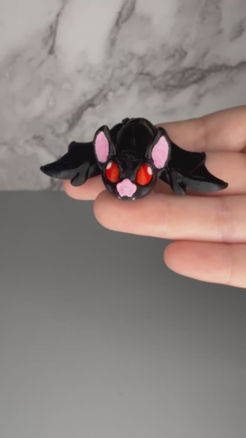 Black bat-shaped figure with red eyes and pink ears on a gray background being shaken to demonstrate articulation